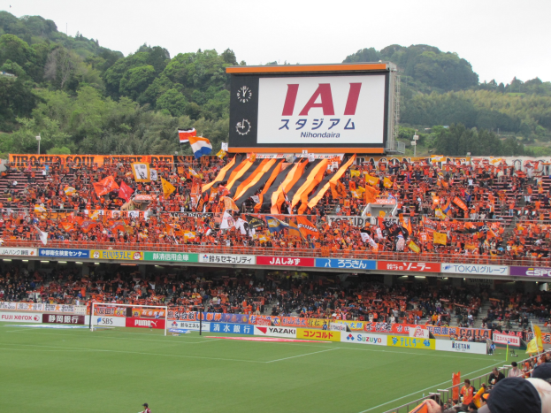Fußballspiel in einem Stadion mit großer Zuschauermenge, saftigem Grün, Torstangen, Bannern, Fahnen, einem großen Bildschirm, Bäumen und einem klaren blauen Himmel.