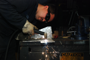 Ein Schweißer in Schutzbrille und Handschuhen bedient eine Schweißmaschine an einem Metallstück, mit einer Warntafel und verstreuten Gegenständen im Hintergrund.