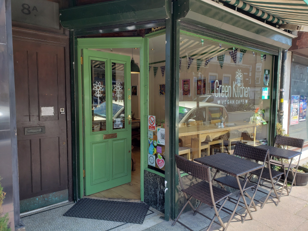 Ein veganes Café namens Green Kitchen Vegan Cafe mit Außenbereich, Topfpflanzen, einer Fußmatte, einer Glastür und einer mit Postern bedeckten Glaswand; drinnen sind Tische, Stühle und eine Hängelampe zu sehen.