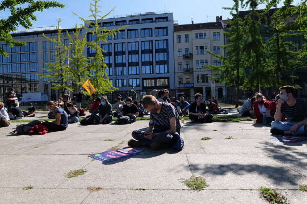 Eine Gruppe von Menschen sitzt vor einem Gebäude auf dem Boden während einer Demonstration in Berlin, einige tragen Masken, mit verstreuten Gegenständen und Bäumen im Hintergrund unter einem klaren blauen Himmel.
