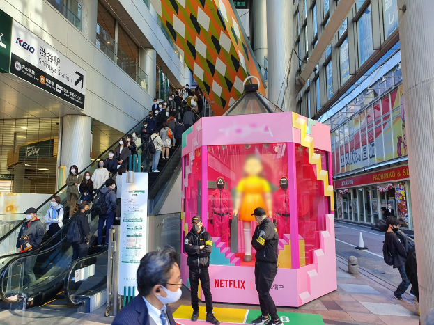 Eine Gruppe von Menschen steht vor einem Gebäude mit Glaswänden, Säulen und Texttafeln, einige tragen Masken, während eine Rolltreppe mit Menschen zu sehen ist. In der Mitte steht eine pinkfarbene Box mit Schaufensterpuppen und der Aufschrift "Netflix's new Netflix booth in Tokyo."
