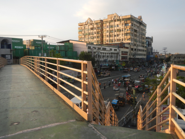 Eine Fußgängerbrücke mit Geländern überspannt eine vielbefahrene Straße mit Fahrzeugen und Fußgängern, mit Gebäuden, Bäumen, Strommasten und einem bewölkten Himmel im Hintergrund.