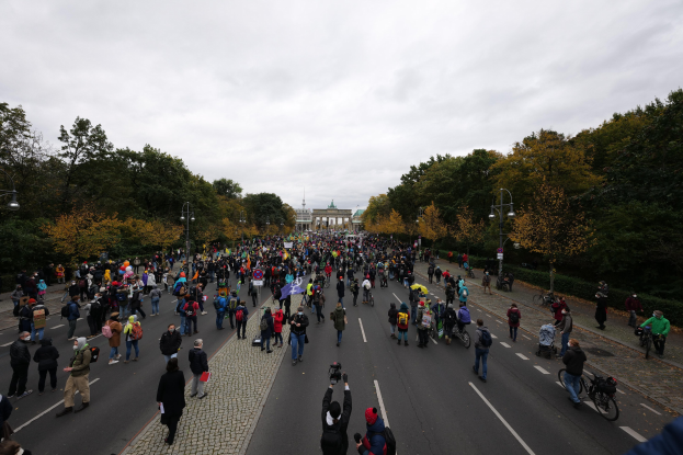 Eine große Gruppe von Menschen marschiert eine baumbestandene Straße in Berlin entlang, mit Kameras bewaffnet, mit einem Gebäude und einem klaren Himmel im Hintergrund.