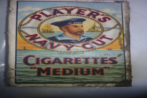 Plakat mit einer Person in einer marineblauen Uniform mit weißem Hemd und roter Krawatte, die Arme verschränkt und einen strengen Gesichtsausdruck hat, bewirbt "Player's Navy Cut Cigarettes Medium" in fetter schwarzer Schrift innerhalb eines weißen Randes.