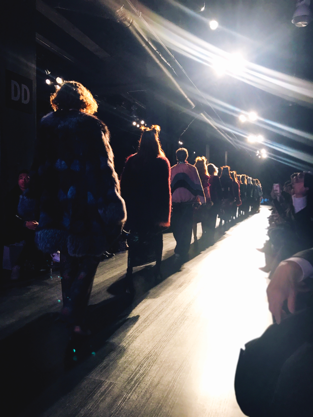 Eine Gruppe von Menschen läuft über den Laufsteg einer Modenschau, beleuchtet von hellen Scheinwerfern, während Zuschauer auf beiden Seiten die Show verfolgen.