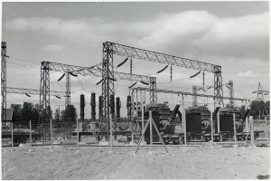 Ein Schwarz-Weiß-Foto eines elektrischen Umspannwerks mit Transformatoren, Masten, Drähten, Maschinen, einem Zaun, Sand, Bäumen und einem bewölkten Himmel, mit Text unten.
