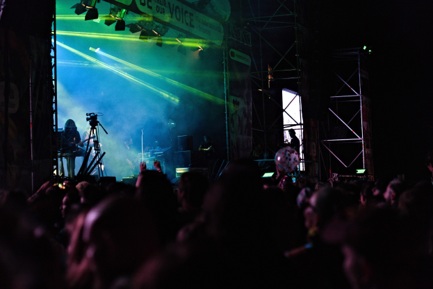 Menschenmenge bei einem Konzert mit Musikern auf der Bühne unter Scheinwerfern, mit einem Banner und Metallrahmen im Hintergrund.