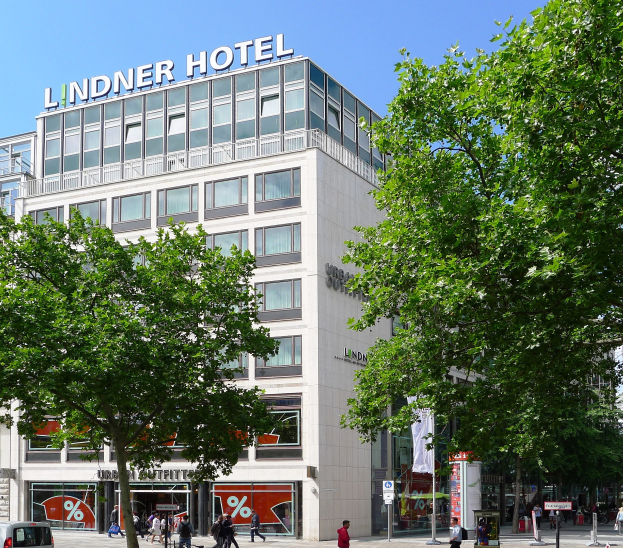 Großes modernes Hotelgebäude mit Glasfenstern und Beschriftung oben, umgeben von Bäumen und einer belebten Straße mit Fußgängern und Fahrzeugen.