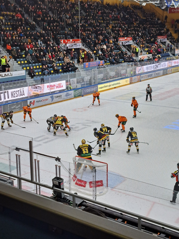 Eine Gruppe von Menschen, die Hockey auf einem Eisring mit einem Netz und einem Zaun spielen, Zuschauer sitzen im Hintergrund und sind von Scheinwerfern beleuchtet.