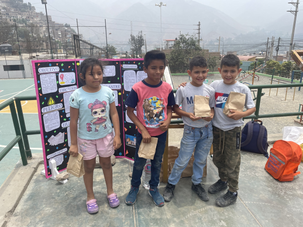 Eine Gruppe von Kindern hält Papiertüten in den Händen und steht auf einem Basketballfeld, rechts davon liegen Tüten und Gegenstände auf dem Boden und links davon eine Tafel mit Text, vor dem Hintergrund von Zäunen, Pfählen, Straßenlaternen, Bäumen, Gebäuden, Hügeln und Himmel.