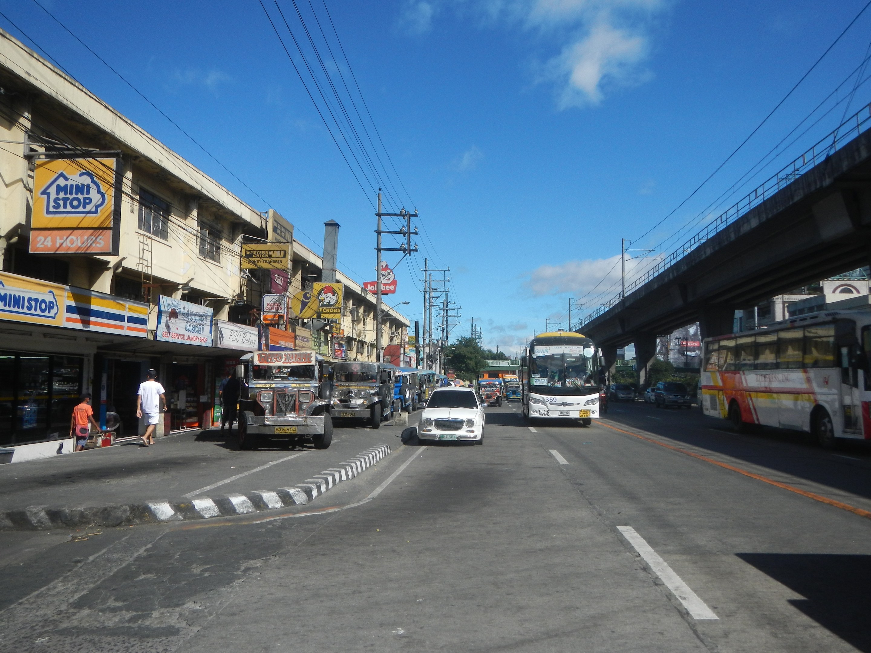 Eine belebte Stadtstraße mit Autos, Bussen, Fußgängern auf dem Gehweg, Gebäuden mit Schildern, Strommasten und -leitungen, einer Brücke, Bäumen und einem bewölkten Himmel.