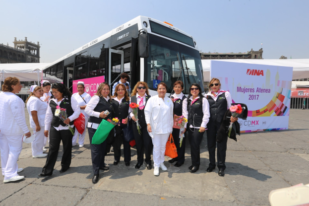 Gruppe von Menschen vor einem Bus mit Blumensträußen, Taschen und einem Banner, Zelten, einem Zaun, einem Gebäude mit Fenstern, einem Fahnenmast mit einer Flagge und einem bewölkten Himmel im Hintergrund.