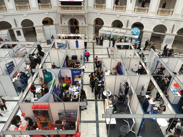 Eine große Gruppe von Menschen auf einer internationalen Fachmesse mit Ständen, Bannern, Tischen, Stühlen und Ballons in einem Raum mit architektonischen Bögen und Säulen.
