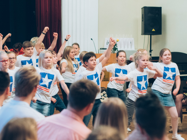 Kinder in weißen Hemden und blauen Shorts tanzen auf einer Bühne mit einem Publikum, das auf Stühlen sitzt, einer Vorhangwand, einem Lautsprecher und einem Tisch mit einem Mikrofon im Hintergrund.