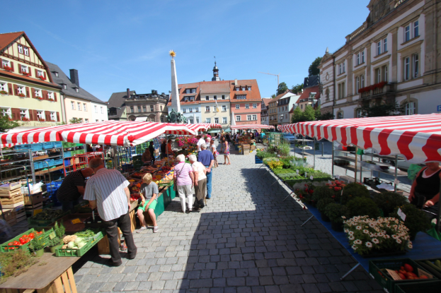 Ein belebter Markt im historischen Stadtzentrum von Heidelberg, Deutschland, mit Menschen, die spazieren gehen, auf Bänken sitzen und in der Nähe von Zelten, Tischen mit Körben voller Gemüse und Gebäuden mit Fenstern im Hintergrund unter einem klaren blauen Himmel.