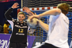 Zwei Männer spielen Handball vor einem Tor mit Netz und Banner im Hintergrund und einigen Zuschauern in einer Stadion.