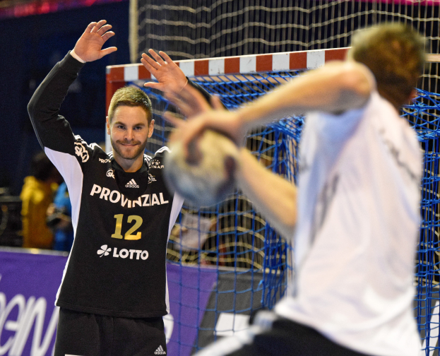 Zwei Männer spielen Handball vor einem Tor mit Netz und Banner im Hintergrund und einigen Zuschauern in einer Stadion.