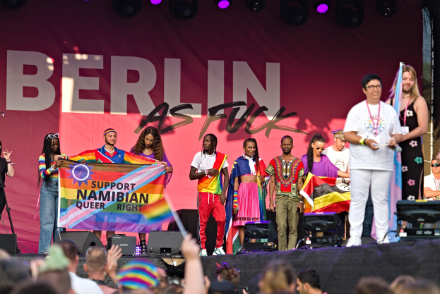 Menschen auf einer Bühne mit einem Banner "Berlin as Fuck" und Lautsprechern, Mikrofonen und einer feiernden Menge unten.