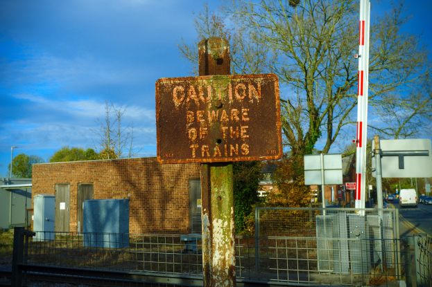 Warnschild an einem Bahnschienenzaun mit Bäumen, Strommästen, einem Gebäude, Containern, Fahrzeugen und einem bewölktem Himmel.