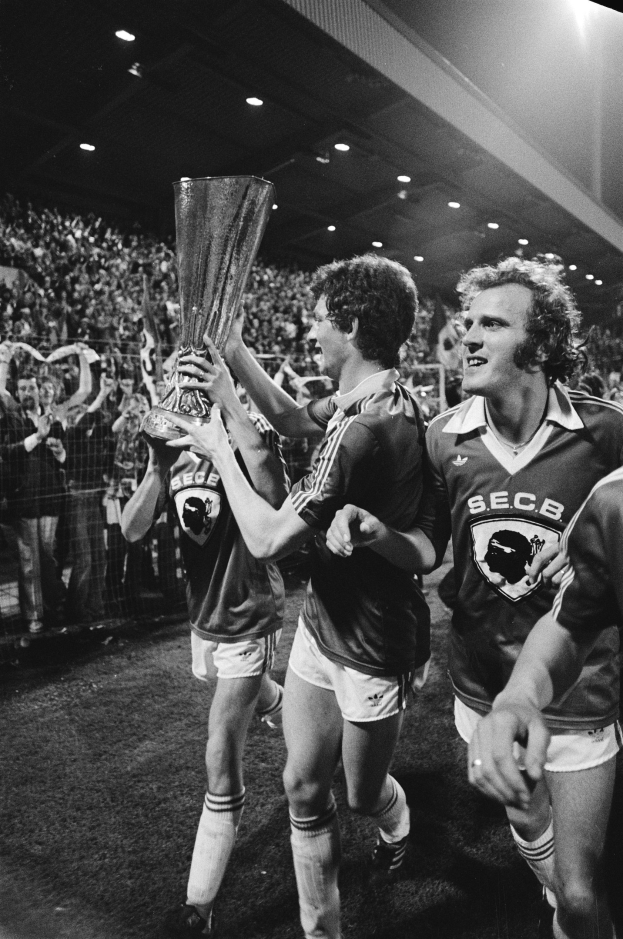 Schwarzes und weißes Bild von Fußballspielern, die mit einem Pokal auf einem Feld stehen und von Zuschauern hinter einem Zaun und Scheinwerfern von oben umgeben sind.