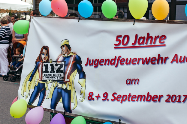 Gruppe von Menschen auf einer Straße mit Kinderwagen, mit einem Banner mit der Aufschrift "50 Jahre Jugendfeuerwehr August 2017", umgeben von Luftballons, einem Zelt, einem Zaun, Bäumen und einem Gebäude mit Fenstern im Hintergrund.