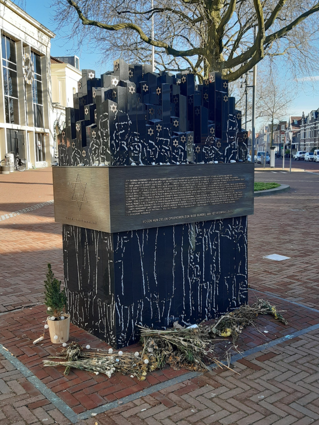 Denkmal in einem Stadtplatz für Holocaust-Opfer, mit einem Baum davor, einem Topfgewächse auf dem Boden, verstreuten trockenen Blättern und Gebäuden, Fahrzeugen, Masten und Bäumen im Hintergrund.