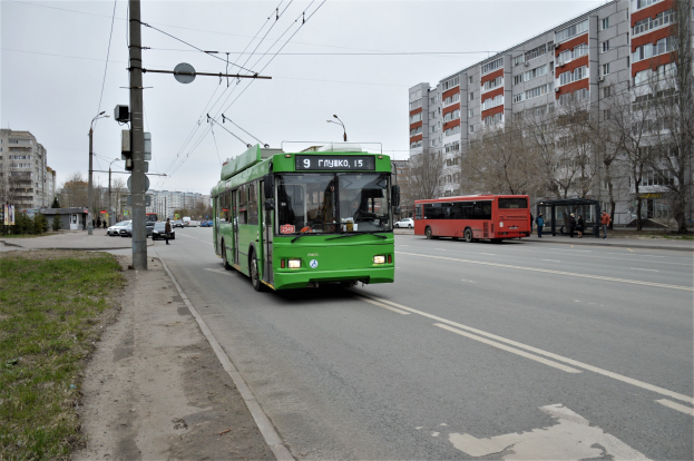 Ein grüner Bus fährt auf einer von hohen Gebäuden gesäumten Stadtstraße, flankiert von Gras, Strommasten und Drähten auf der linken Seite und Bäumen und einem blauen Himmel im Hintergrund.