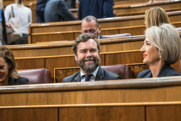 Eine Gruppe von Menschen, die in einem Gerichtssaal sitzen, mit einem Mann in Anzug und zwei Frauen in legerer Kleidung nebeneinander, Bänke sind im Hintergrund zu sehen.