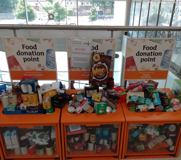 Orangene Schränke voller Lebensmittelspenden, darunter Flaschen, Dosen und Kartons, mit Informationsschildern, durch Glasfenster mit Blick auf Fahrzeuge, Bäume, Gebäude und Himmel.