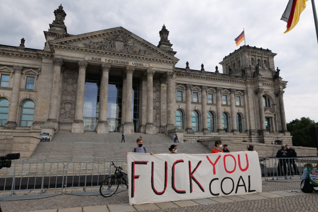 Gruppe von Menschen mit einem "Fuck You Coal"-Plakat vor dem Reichstaggebäude in Berlin, Deutschland, mit Bäumen, Fahnenmast und bewölktem Himmel im Hintergrund.