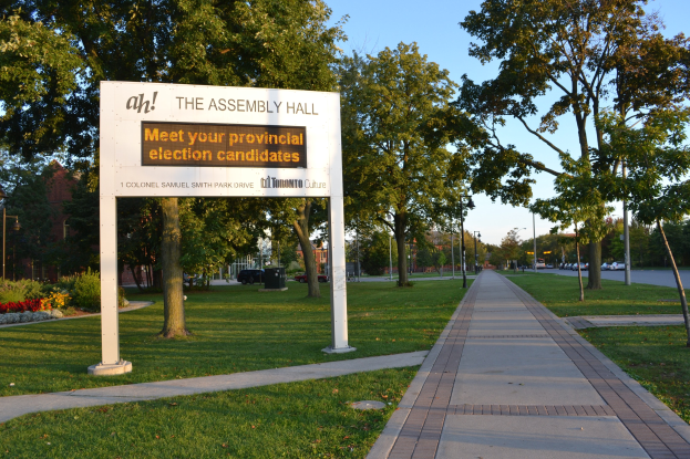 Vorstand auf Gras mit Text "Die Versammlungshalle - Treffen Sie Ihre Landtagswahlkandidaten" umgeben von Weg, Bäumen, Pflanzen, Straßenlaternen, Fahrzeugen, einem Gebäude und einem bewölkten Himmel.